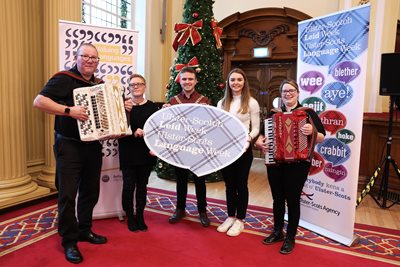 Line up of five people with two holding accordions with banners and a prop promoting Ulster-Scots Language Week
